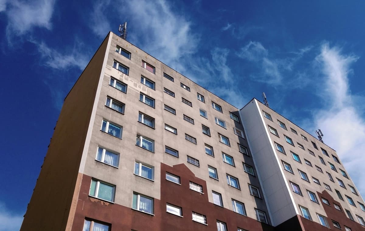 Ett flerbostadshus med en grå och brun fasad under en blå himmel med vita moln.